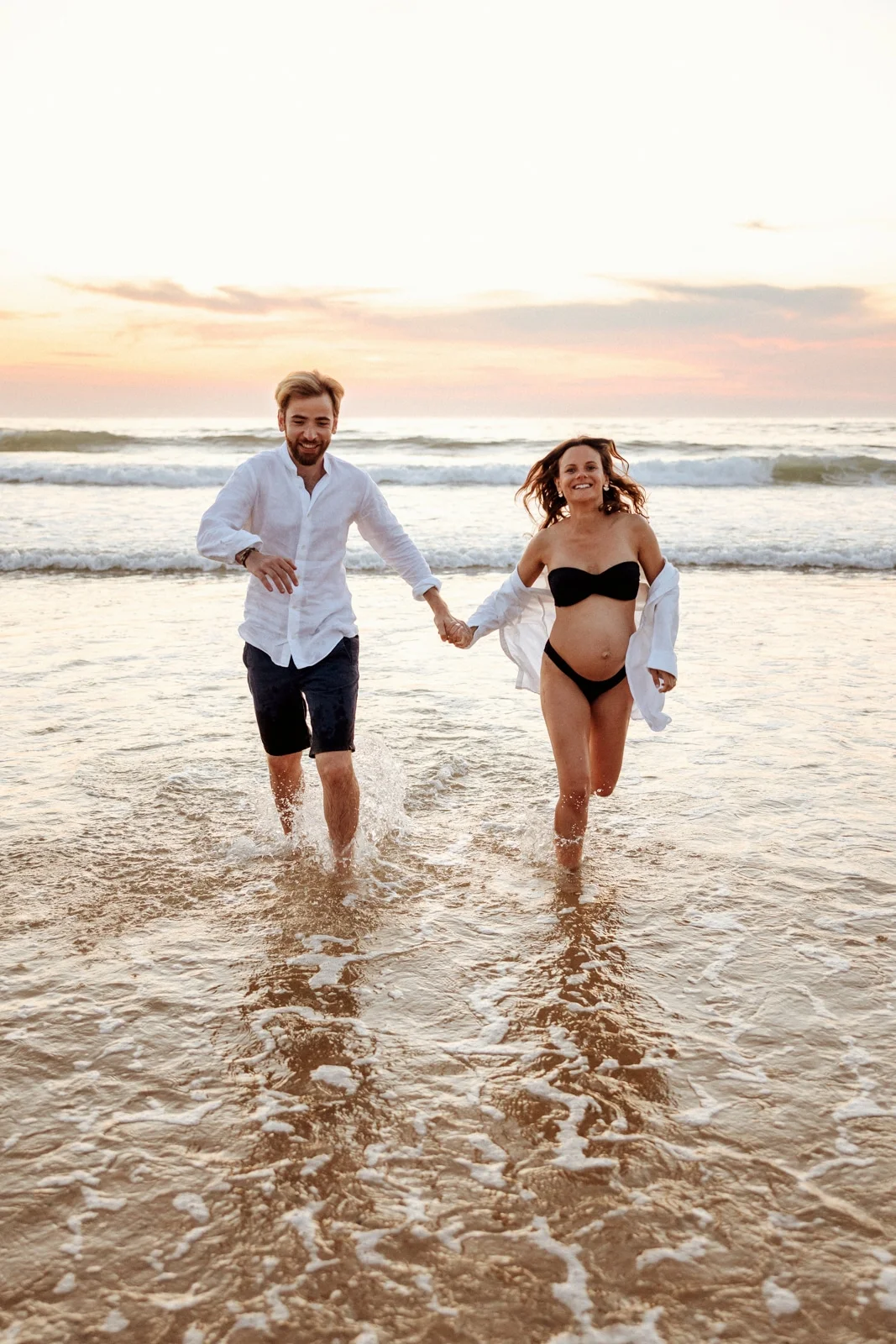 Couple attendant un bébé courant dans l’eau lors d’une séance grossesse sur la plage dans les Landes au coucher du soleil