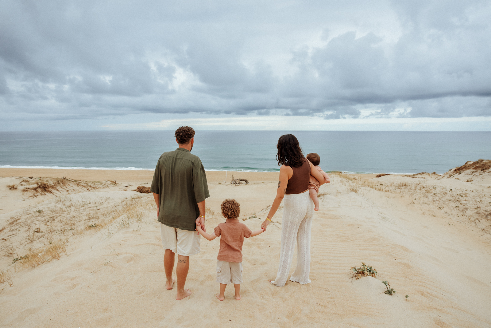 Une famille de quatre personnes marche dans les dunes vers l'océan, les parents tenant leurs enfants par la main ou dans les bras.