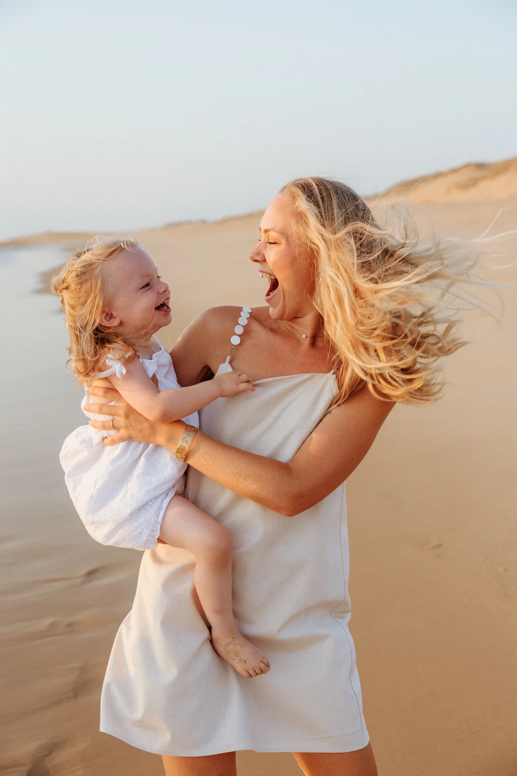 Une maman et sa petite fille éclatent de rire ensemble sur la plage, photographiées dans la lumière douce de fin de journée.